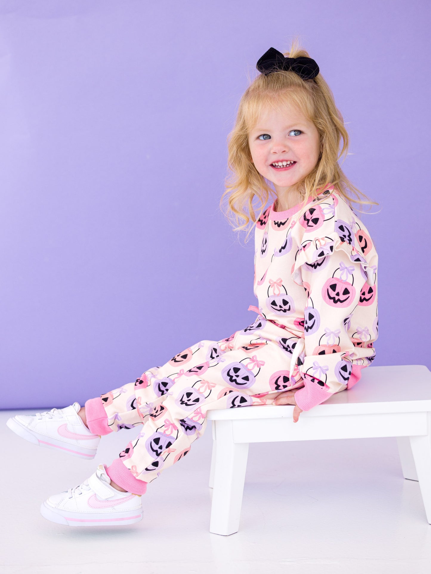 Child wearing a Halloween-themed jogger set with pumpkins and ghosts, sitting on a white stool against a purple background.