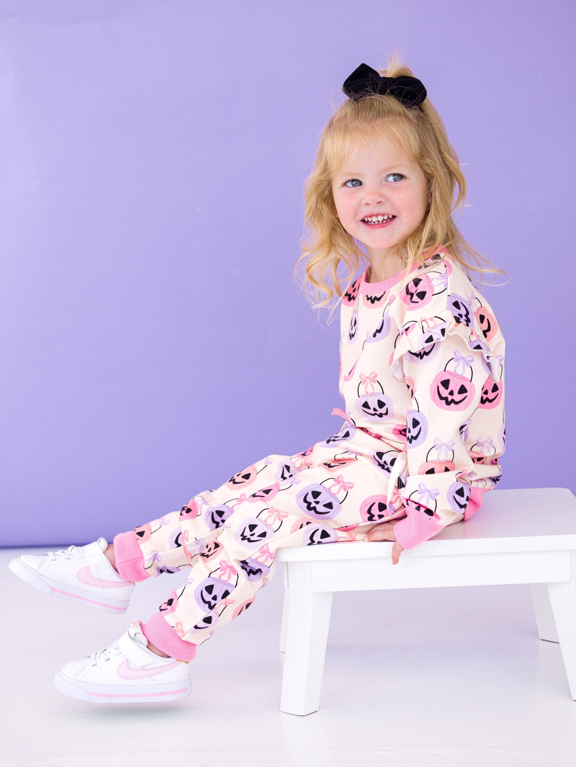 Child wearing a Halloween-themed jogger set with pumpkins and ghosts, sitting on a white stool against a purple background.
