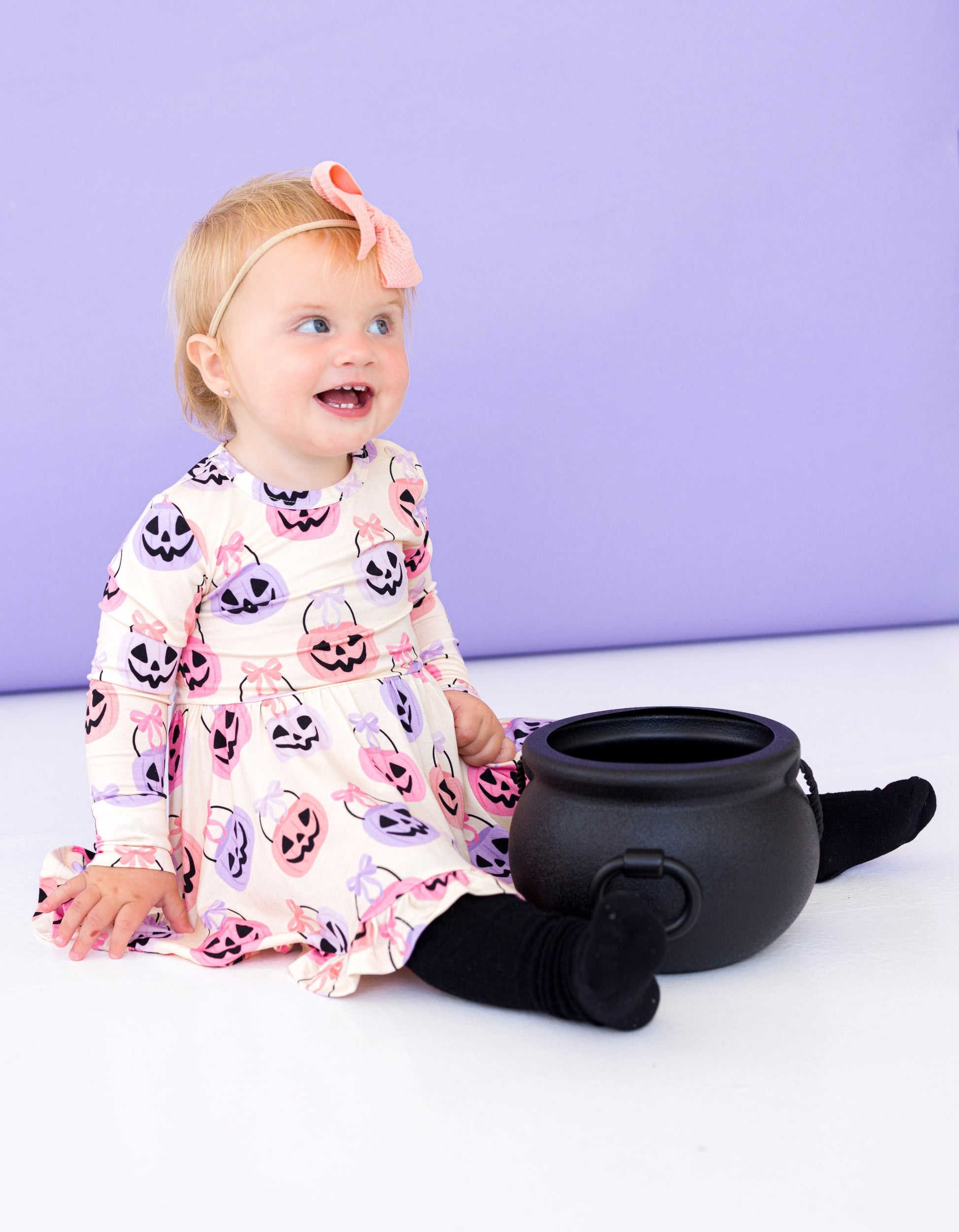 Child wearing a bamboo Halloween-themed dress with pumpkins and ghosts, sitting next to a black cauldron on a white surface with a purple background.