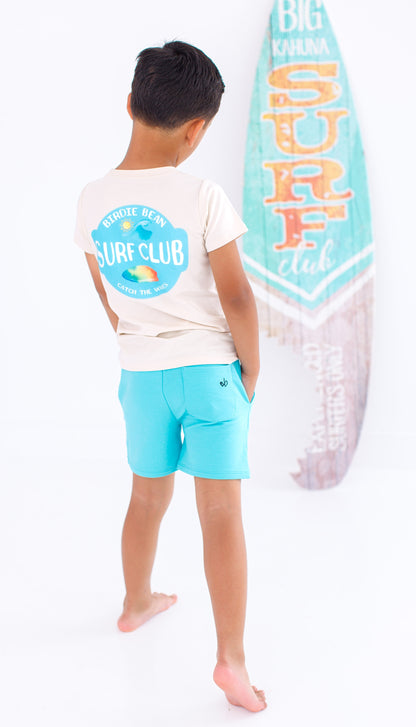 Child wearing a beige graphic t-shirt standing next to a surf board