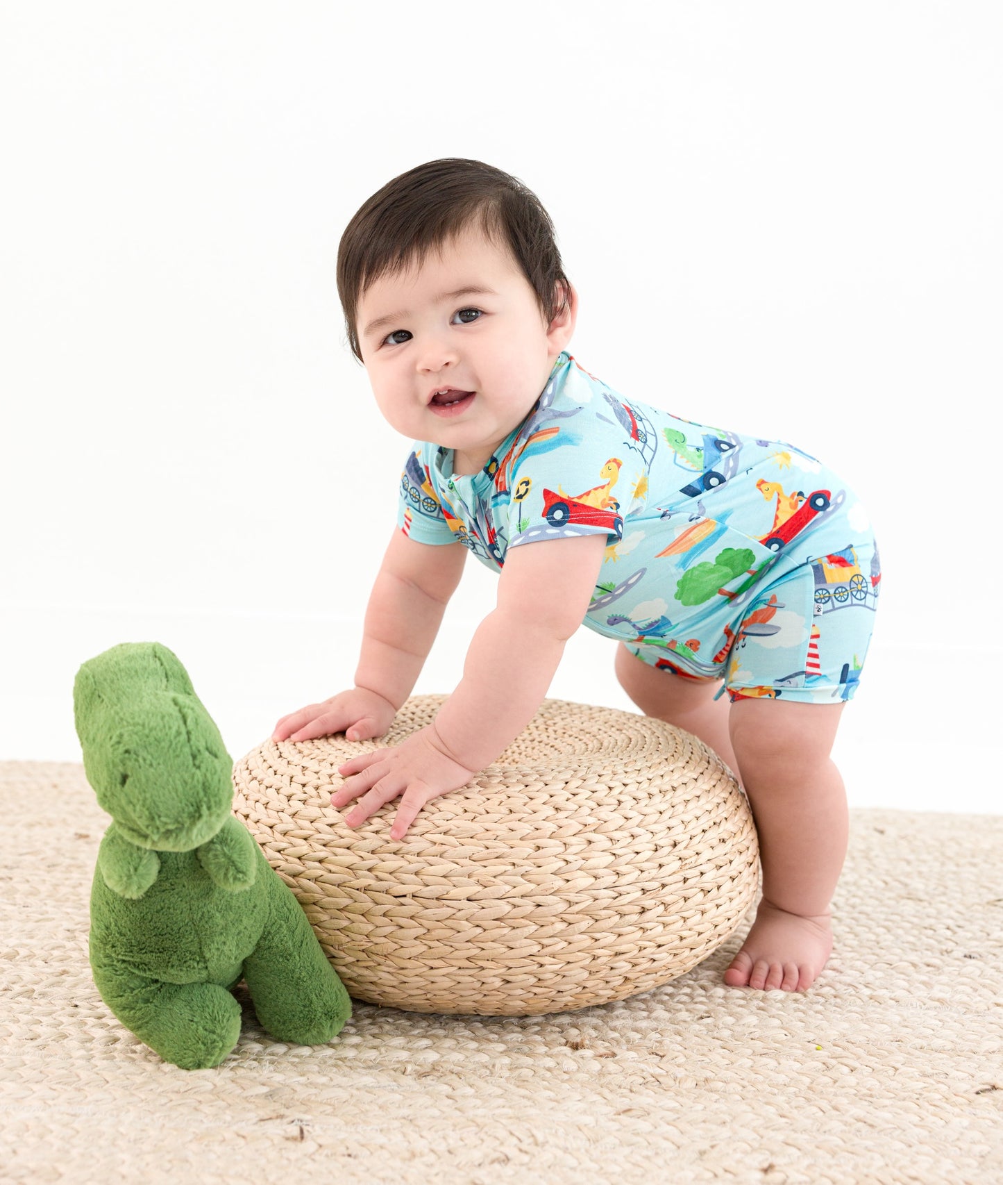 Baby in a colorful bamboo outfit standing next to a woven stool and green dinosaur toy on a white background