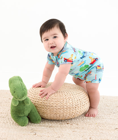 Baby in a colorful bamboo outfit standing next to a woven stool and green dinosaur toy on a white background
