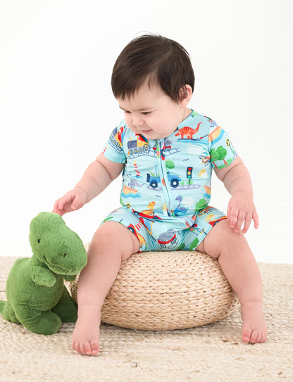 Baby in a colorful bamboo onesie with vehicle pattern sitting on a woven stool next to a green plush toy.