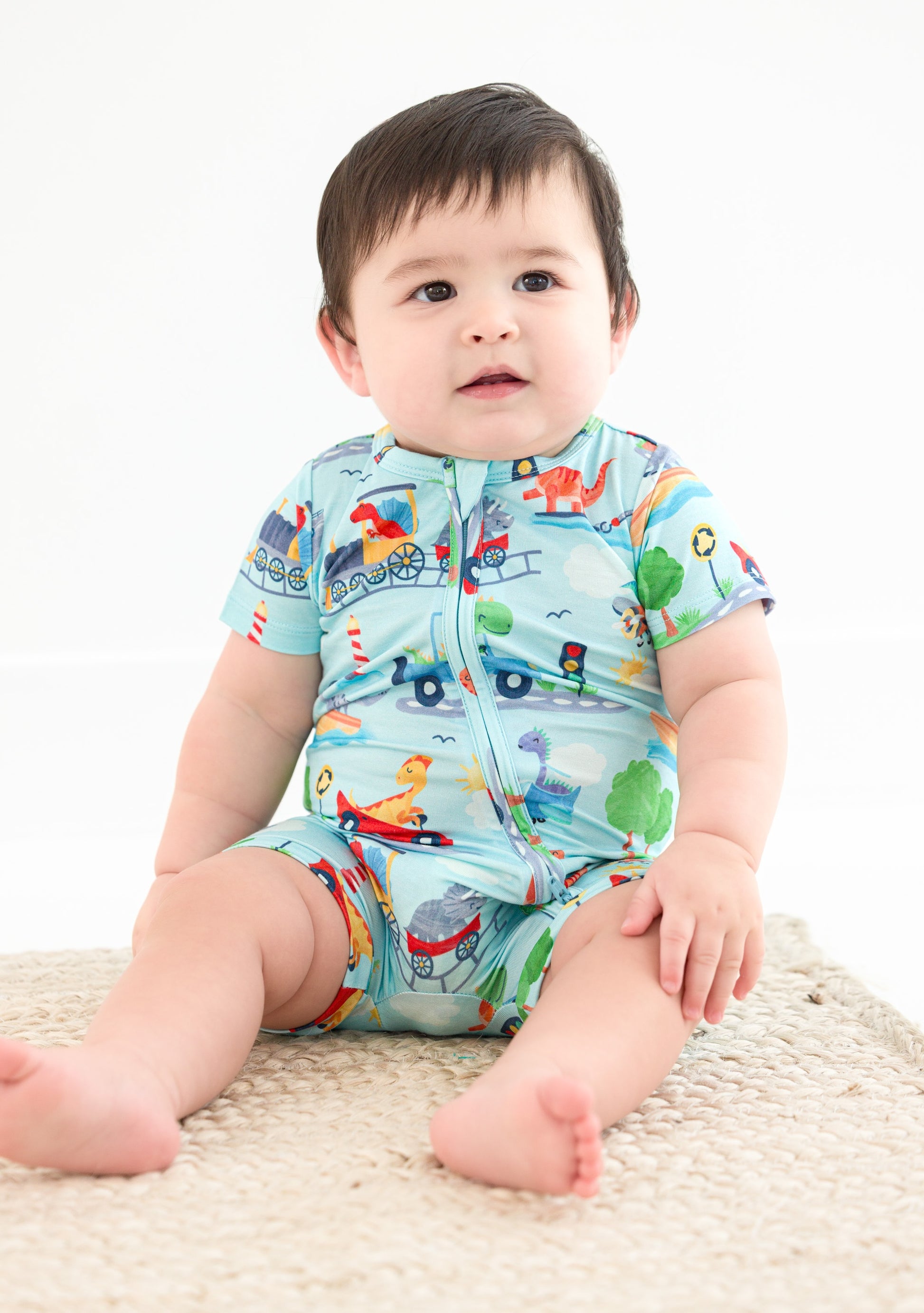 A baby sits, wearing a short-sleeve zip romper. The bamboo zip romper features animals driving trains, boats, cars, etc.