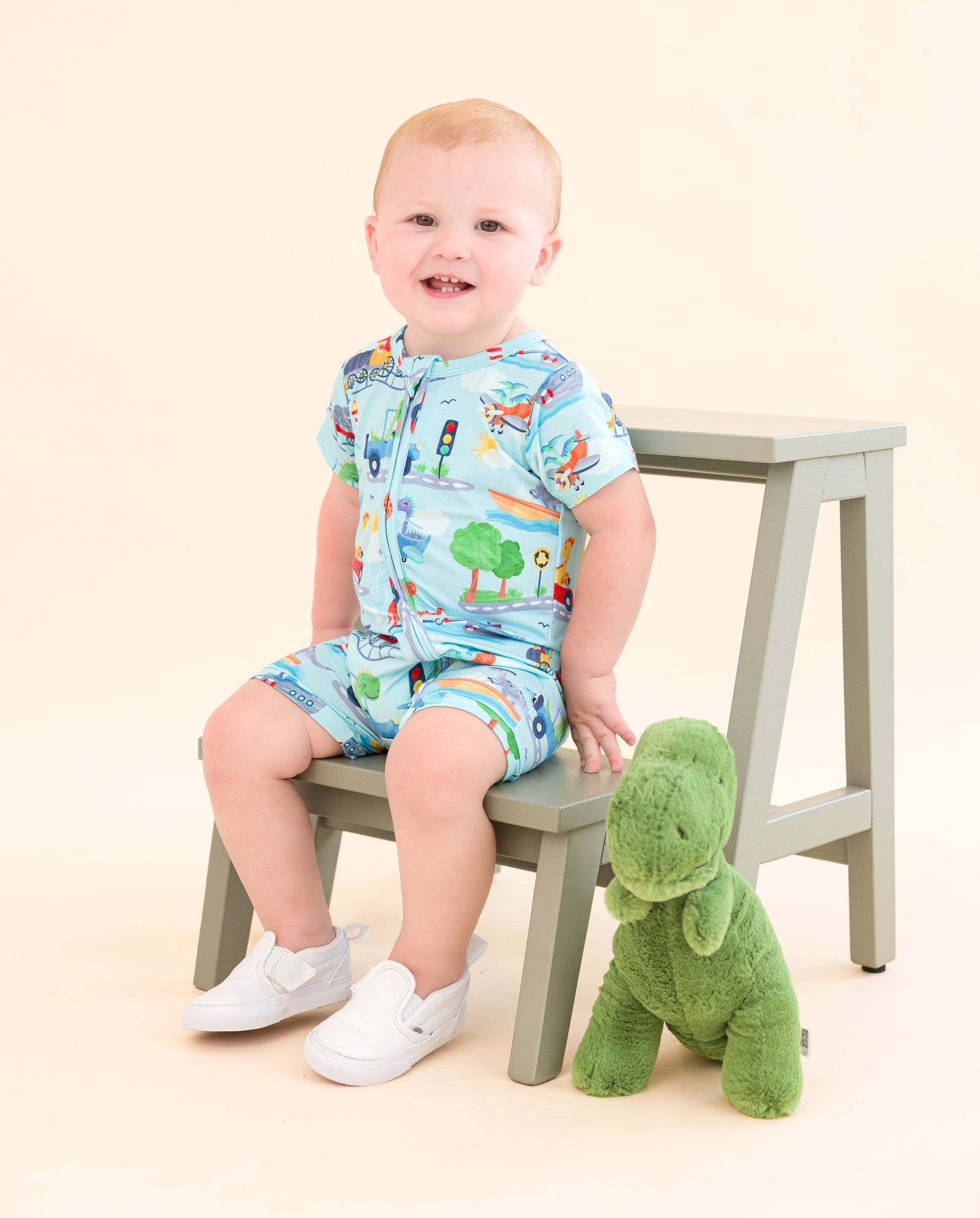 A toddler sits, wearing a short-sleeve zip romper. The bamboo zip romper features animals driving trains, boats, cars, etc.