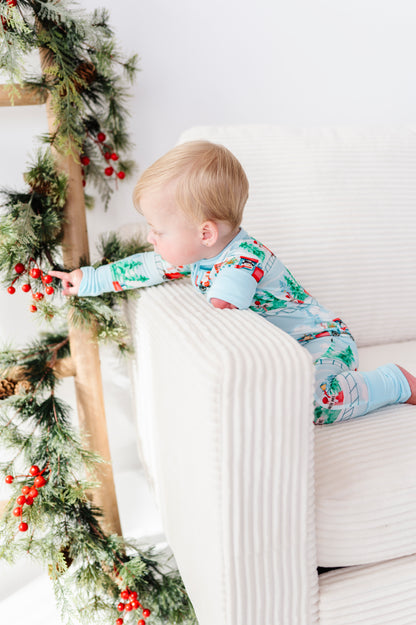 A baby wearing a light blue Christmas-themed bamboo pajama set with a Santa Claus and snowman print, crawling on a white ribbed armchair next to a pine garland with red berries.