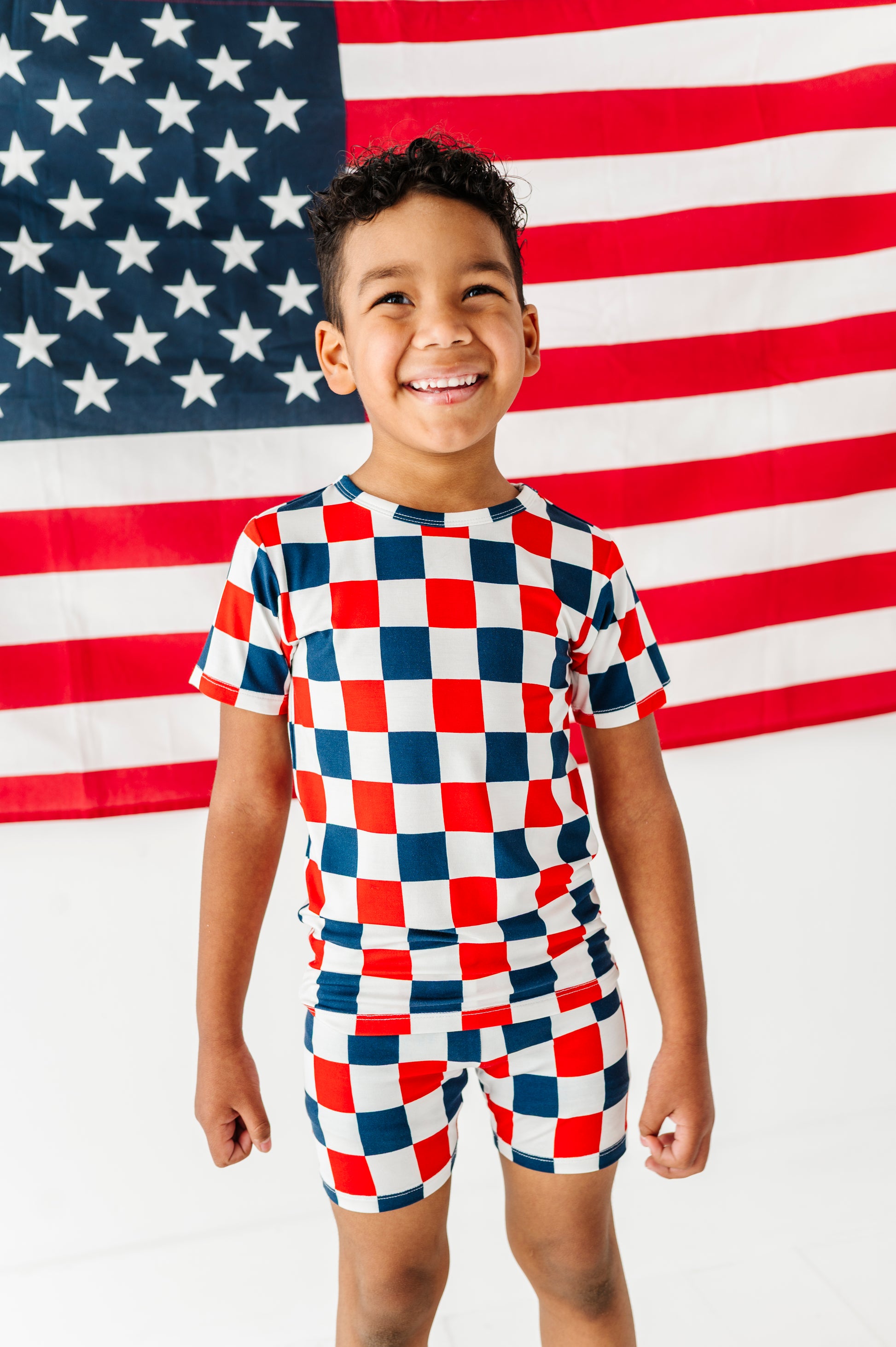 Child wearing a red, white, and blue checkered bamboo pj outfit in front of an American flag.