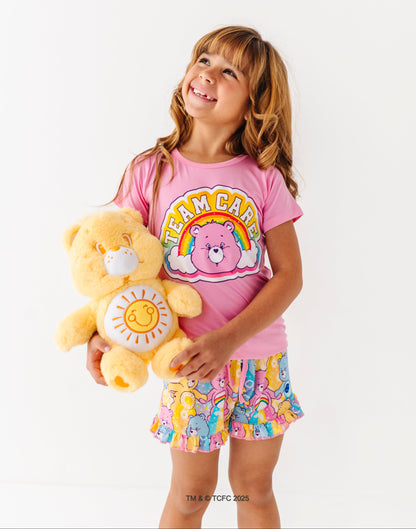 Child holding a yellow Care Bears plush while wearing the pink Care Bears™ Stare pajama top and shorts