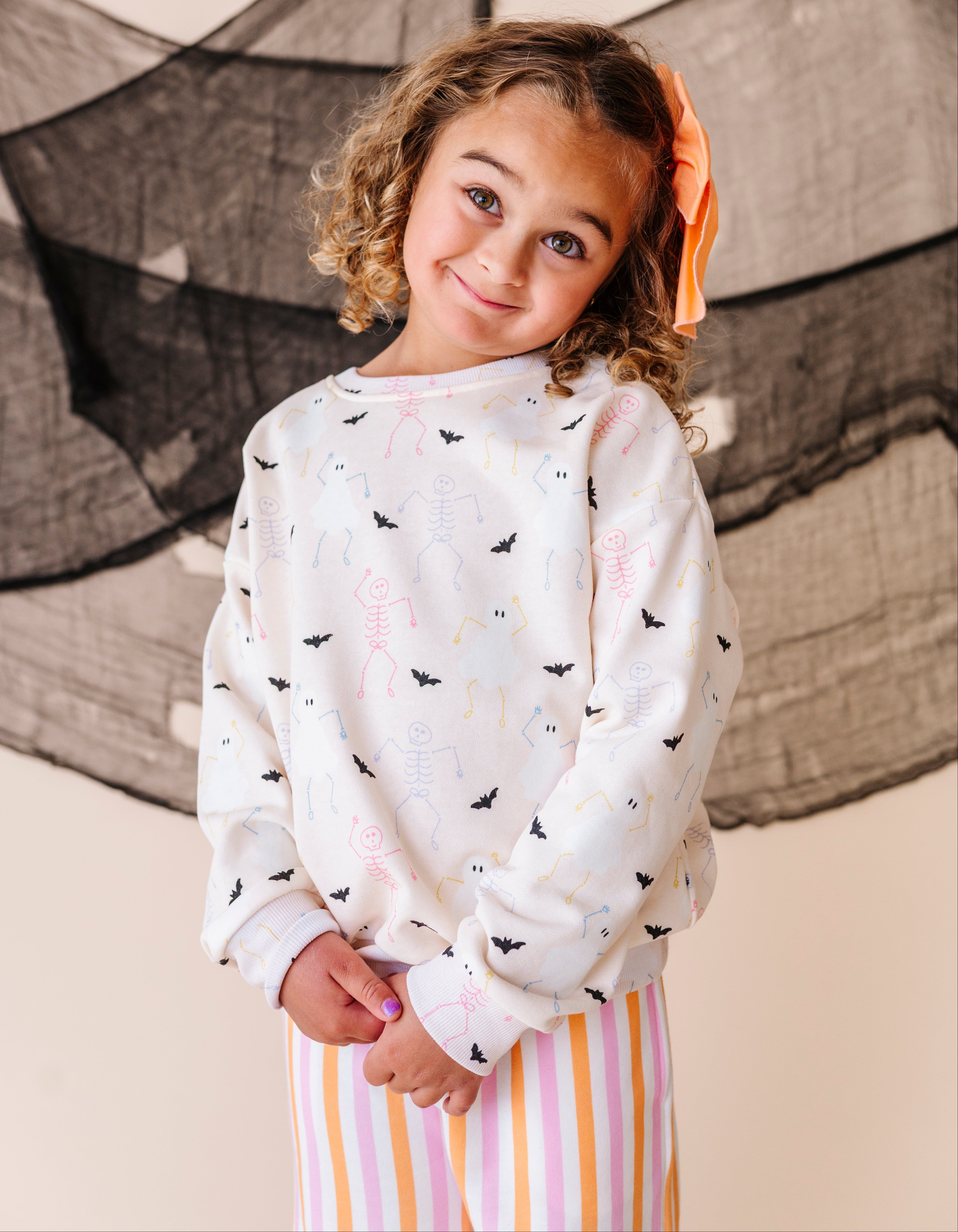 Young girl wearing a white sweatshirt with black and colorful bird patterns, standing in front of a decorative curtain.