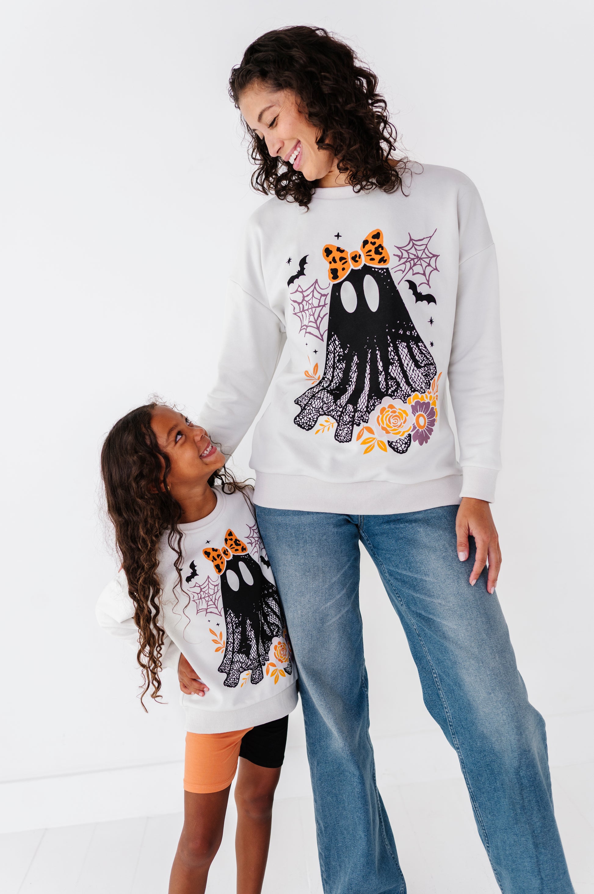 A woman and a child wearing matching white Halloween sweatshirts with a black ghost graphic, orange leopard ears, and spiderweb motifs, paired with blue jeans against a white studio background.