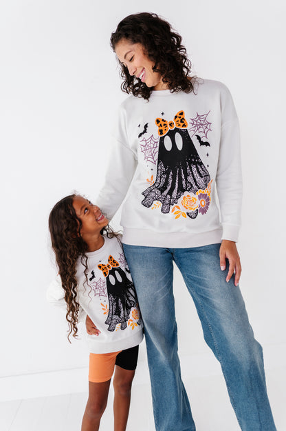 A woman and a child wearing matching white Halloween sweatshirts with a black ghost graphic, orange leopard ears, and spiderweb motifs, paired with blue jeans against a white studio background.