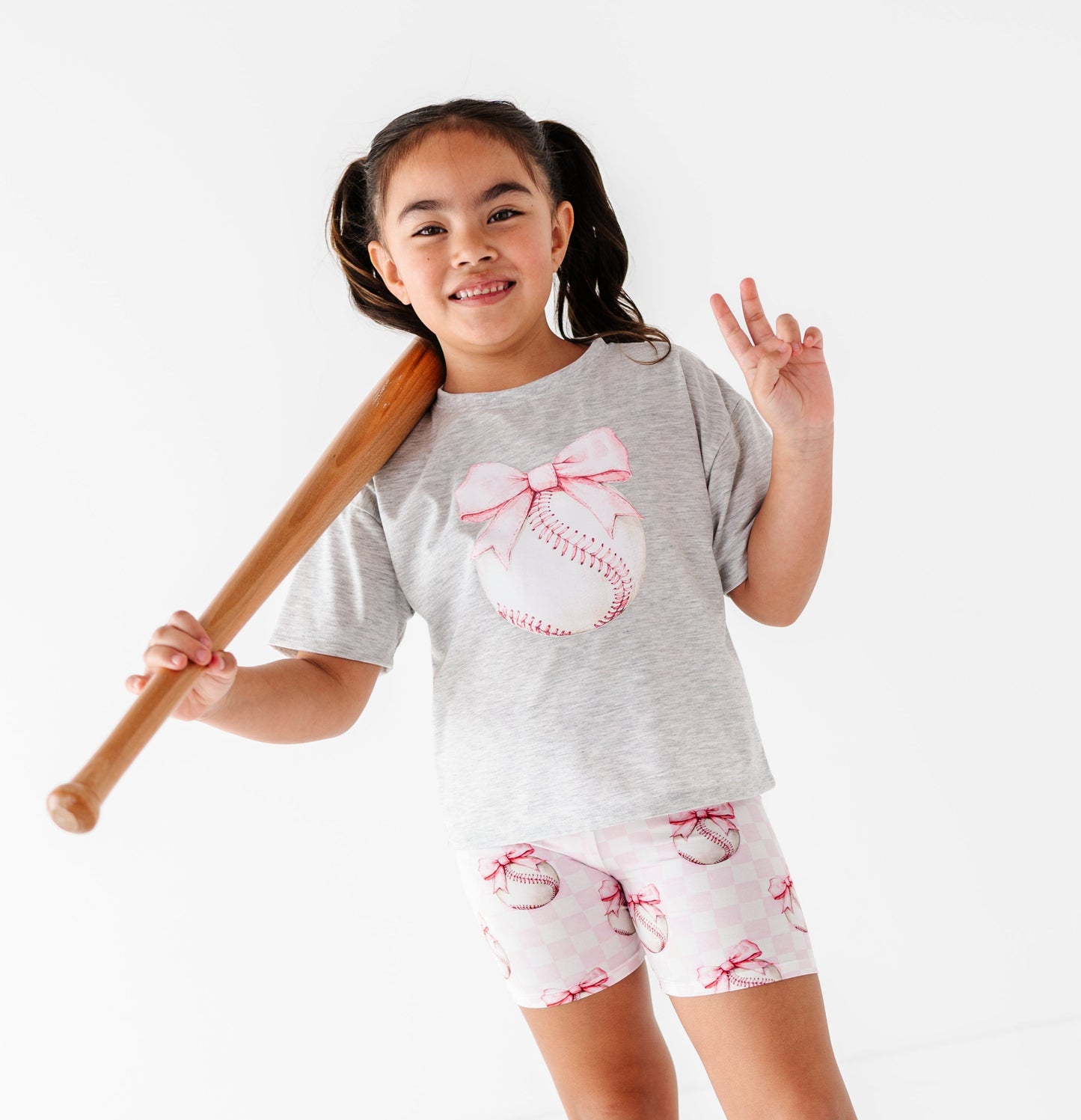 Young girl in gray t-shirt and bamboo biker shorts with bows, holding a baseball bat on a white background