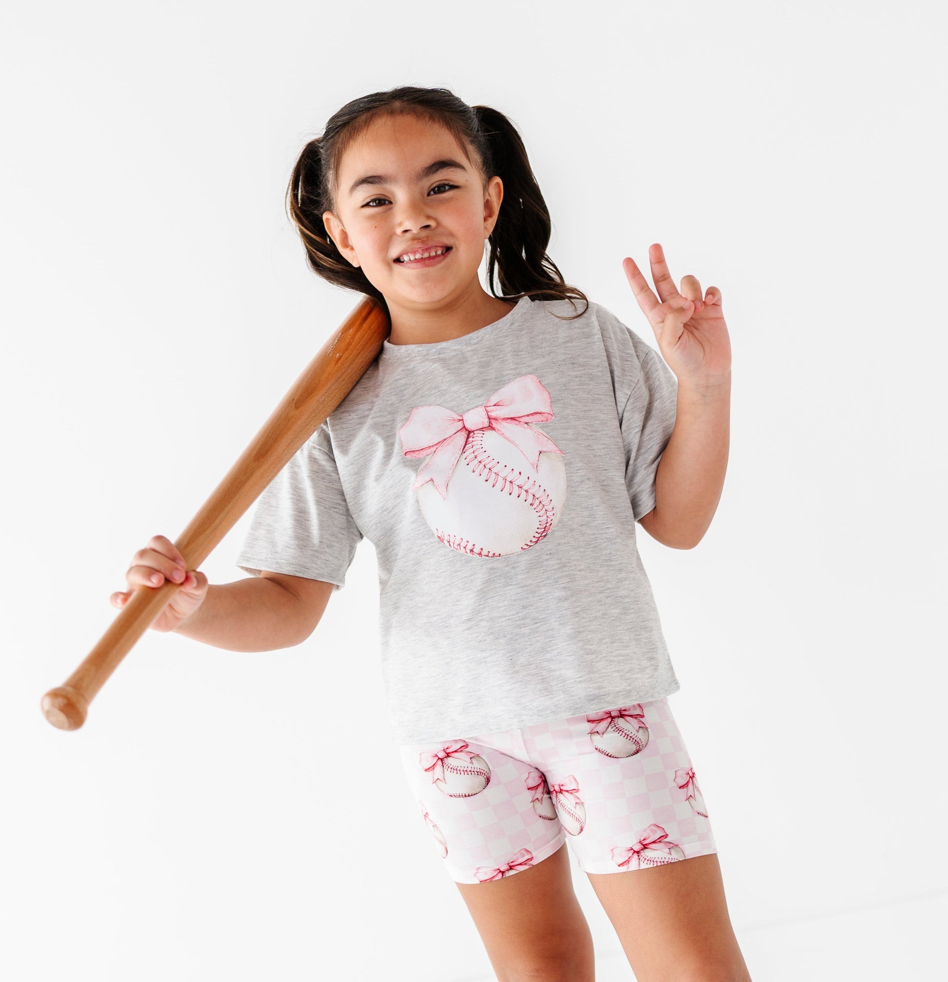 Young girl in gray t-shirt and bamboo biker shorts with bows, holding a baseball bat on a white background