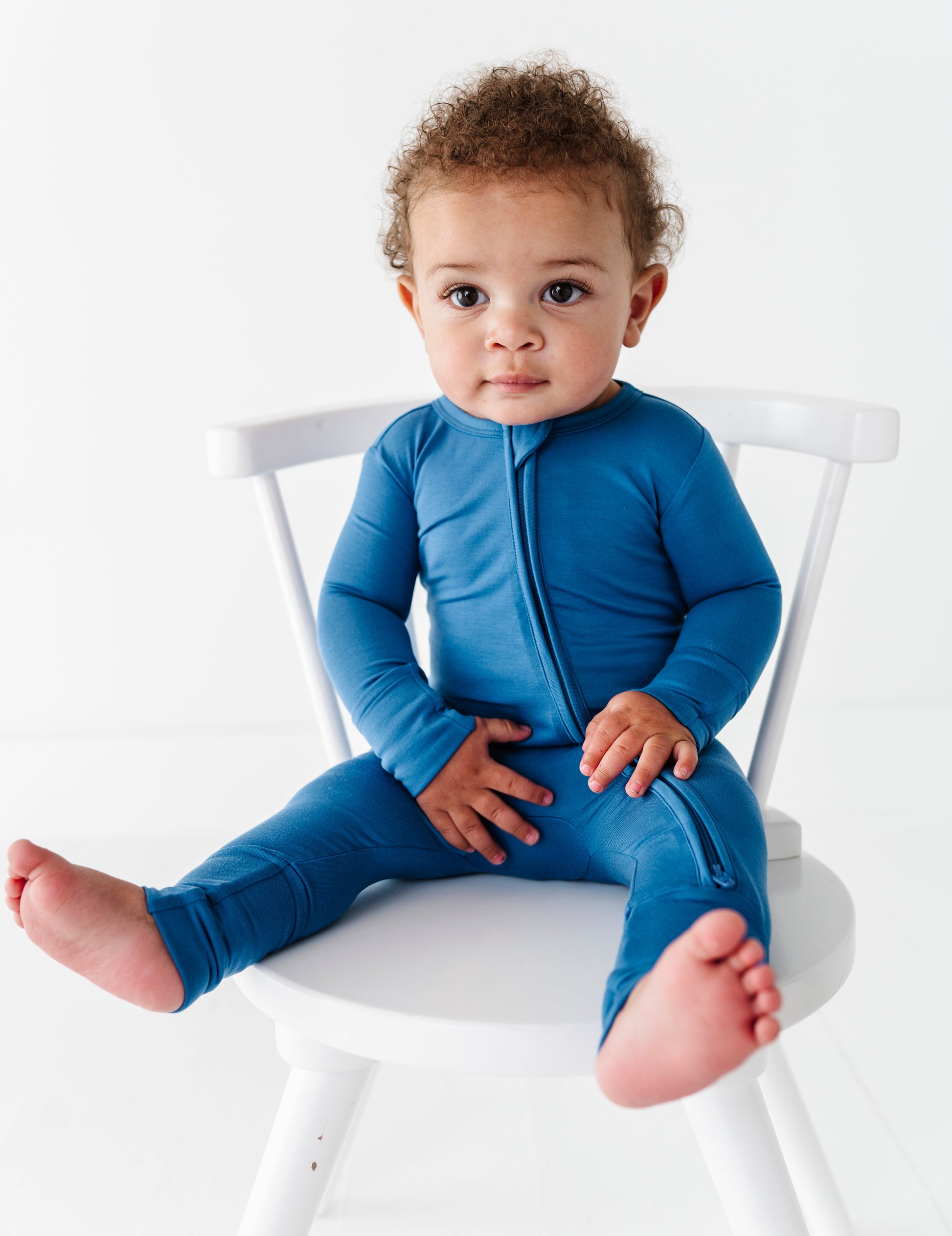 Baby wearing a blue bamboo convertible romper sitting on a white chair against a white background