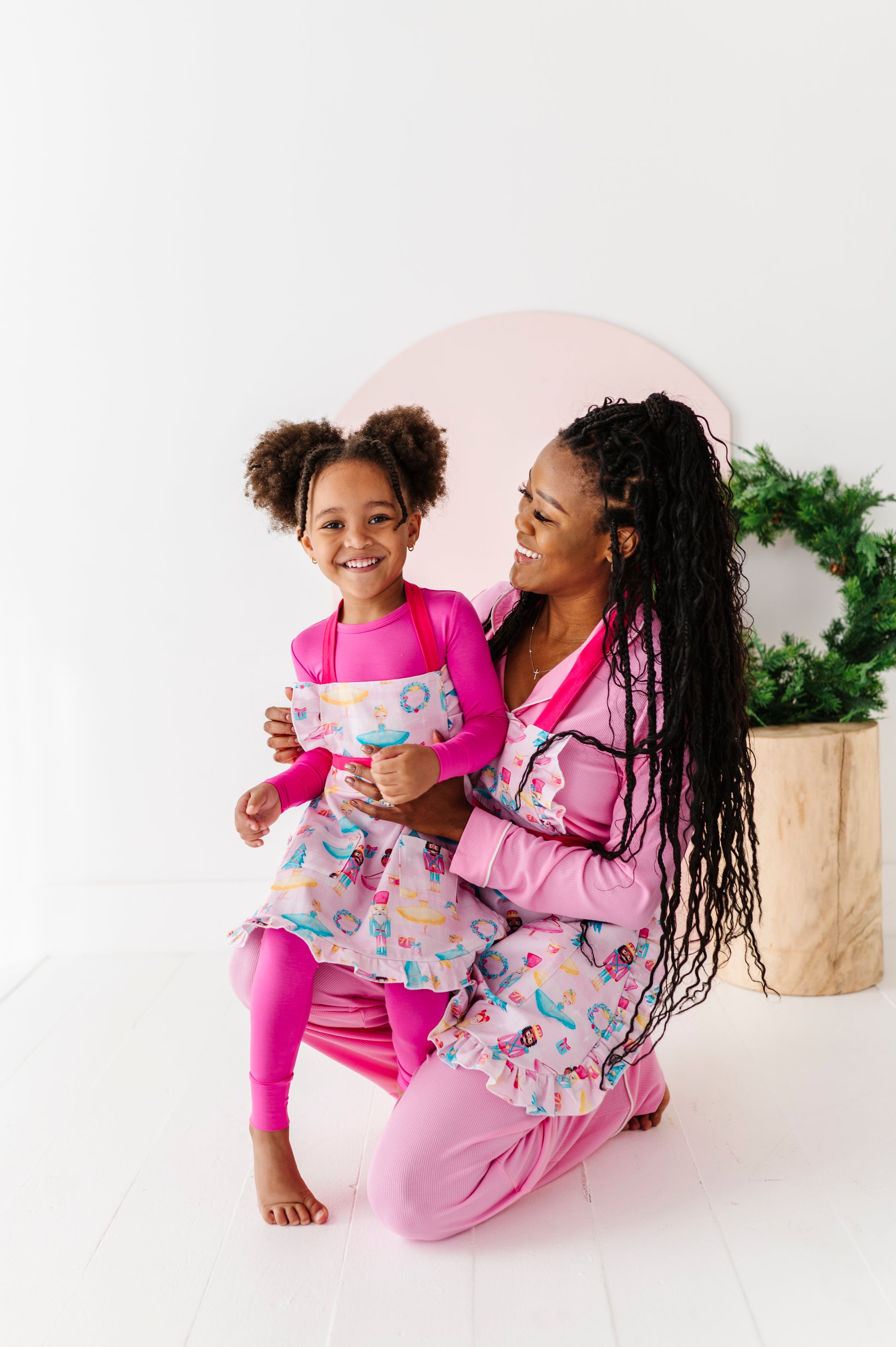 Woman and child in matching light pink aprons featuring ballerinas, nutcrackers, wreaths, presents, and Christmas trees. 
