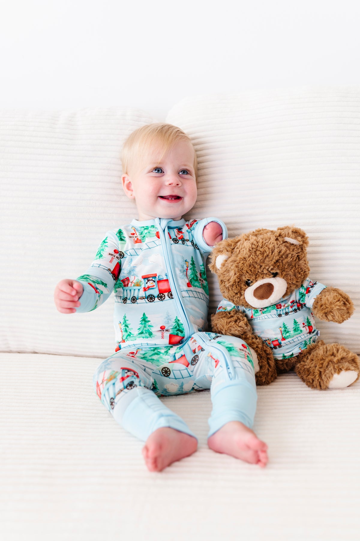 A baby sitting on a white textured couch wearing a light blue long-sleeve bamboo pajama set with a colorful Christmas tree truck pattern, next to a brown teddy bear wearing a matching pajama set.