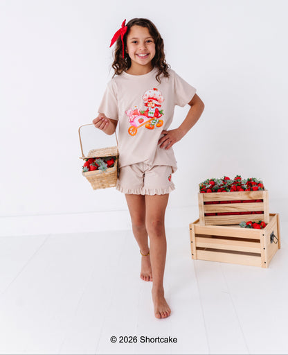 Young girl in a beige Strawberry Shortcake™ outfit with a red bow, holding a basket of strawberries, standing next to wooden crates filled with strawberries on a white background.