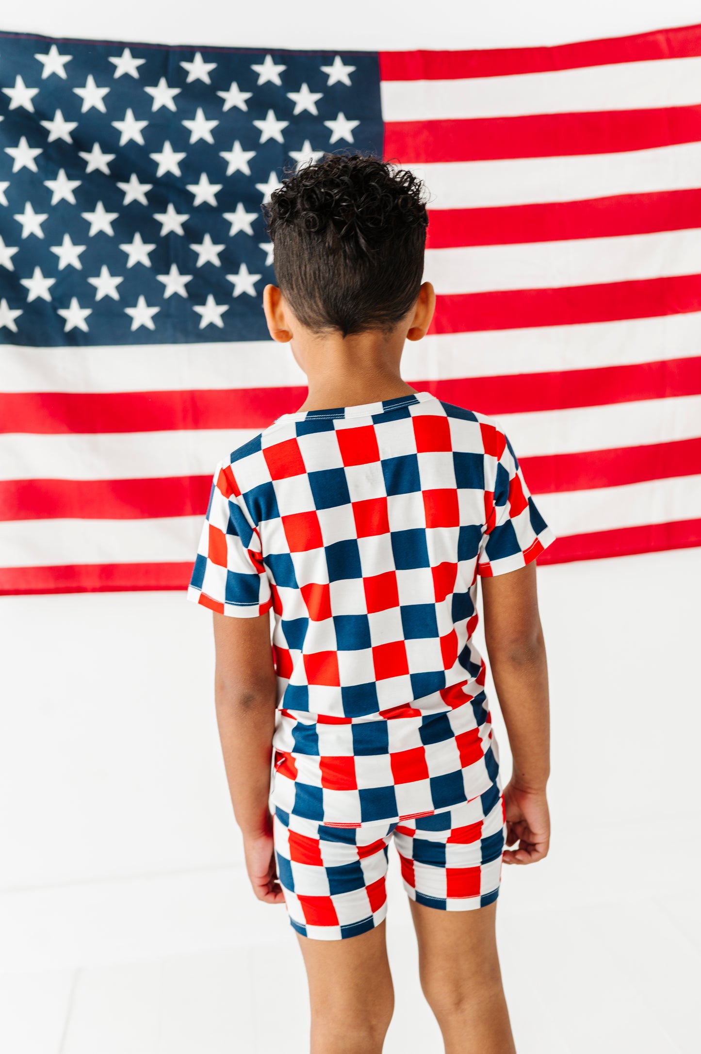 Little boy wearing a red, white, and blue checkered bamboo short set standing in front of an American flag.