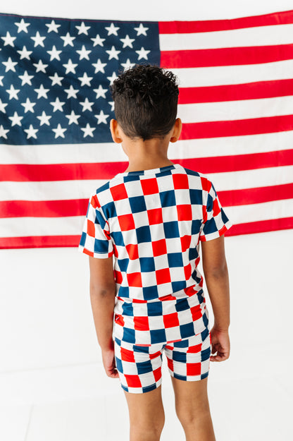 Little boy wearing a red, white, and blue checkered bamboo short set standing in front of an American flag.