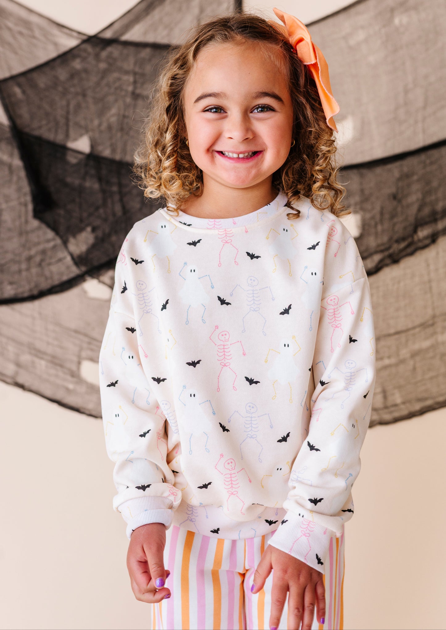 Young girl wearing a cream sweatshirt with black and colorful halloween patterns, standing in front of a decorative net.