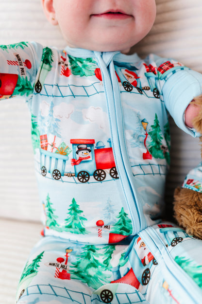 A close-up of a baby wearing a light blue zip-front footed bamboo sleeper with a festive Christmas train print, lying on a white textured blanket.