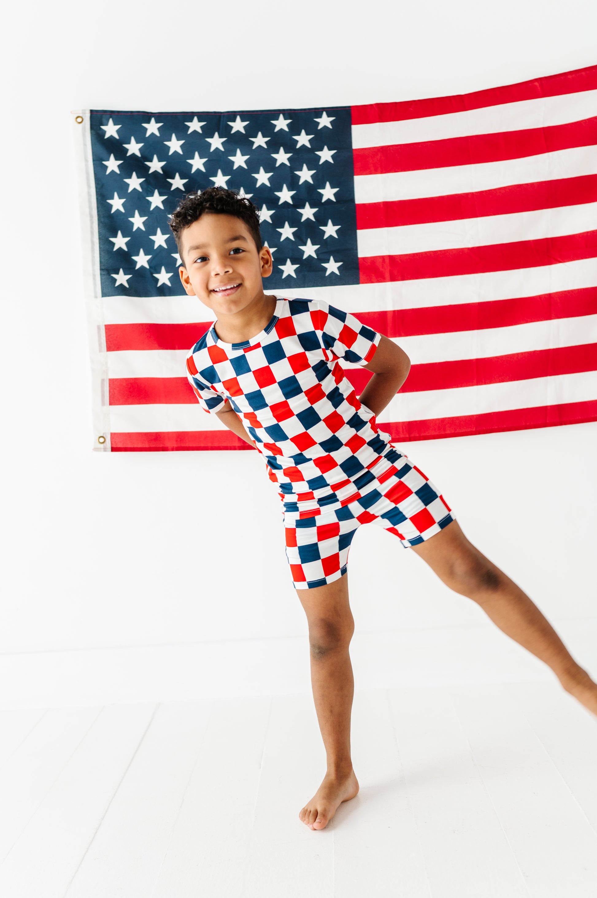 Child wearing a red, white, and blue checkered bamboo pajama outfit standing in front of an American flag.