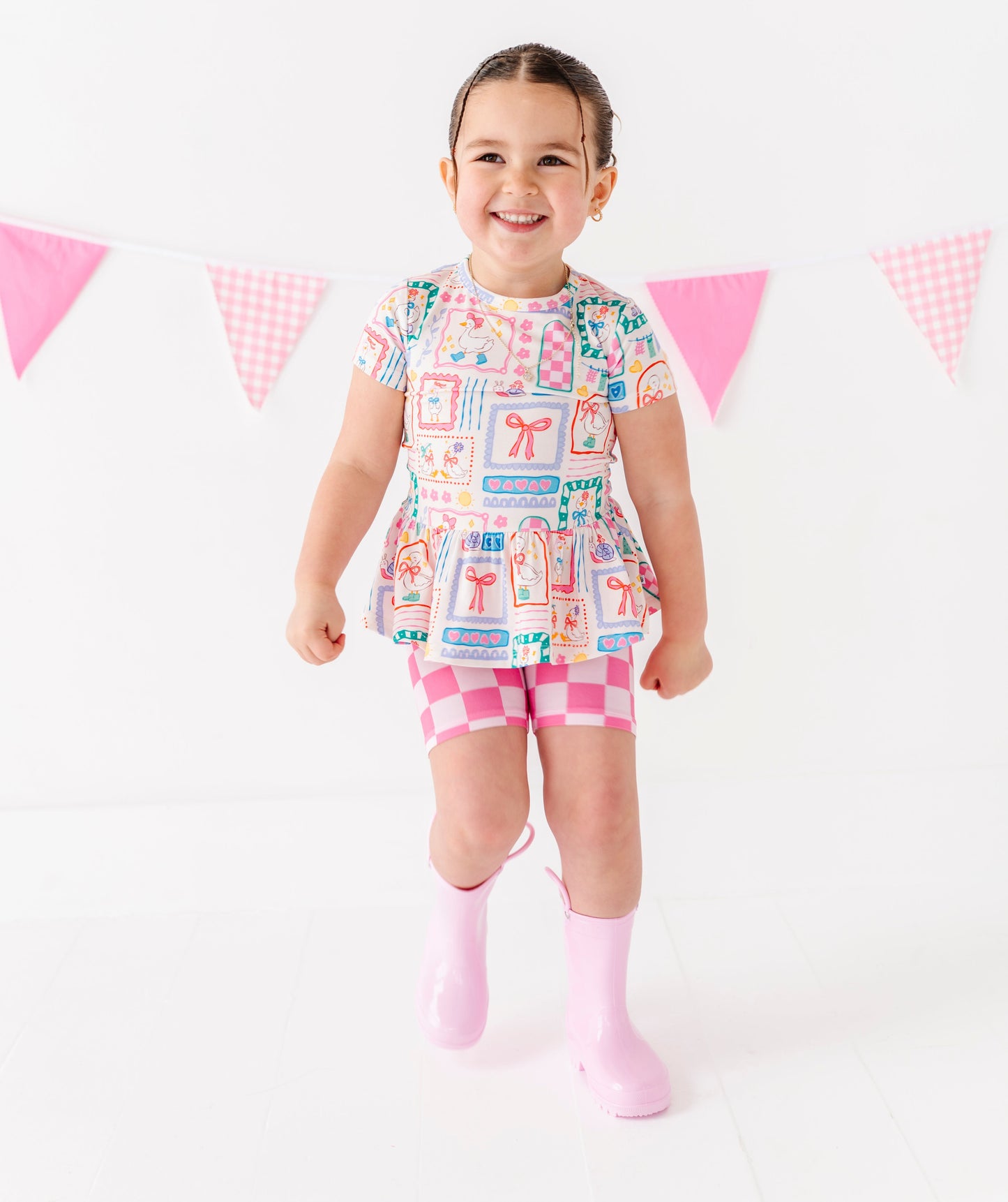 Child wearing a colorful dress with a pattern of boxes and a pink skirt, standing against a white background with pink flags.