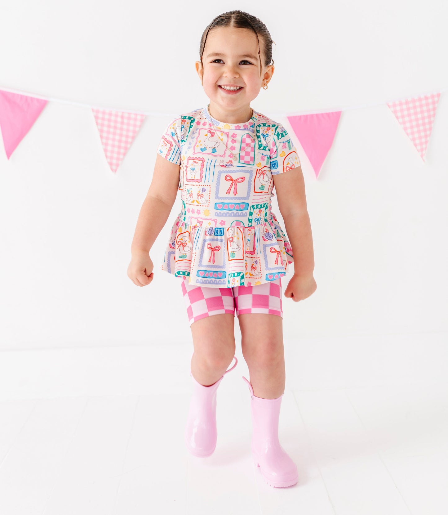 Child wearing a colorful dress with a pattern of boxes and a pink skirt, standing against a white background with pink flags.