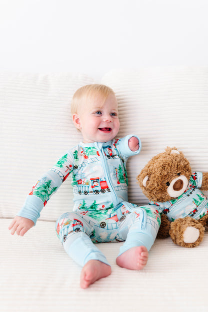 A baby sitting on a white textured blanket next to a brown teddy bear, wearing a light blue long-sleeve bamboo footed pajama with a colorful train and Christmas tree print.