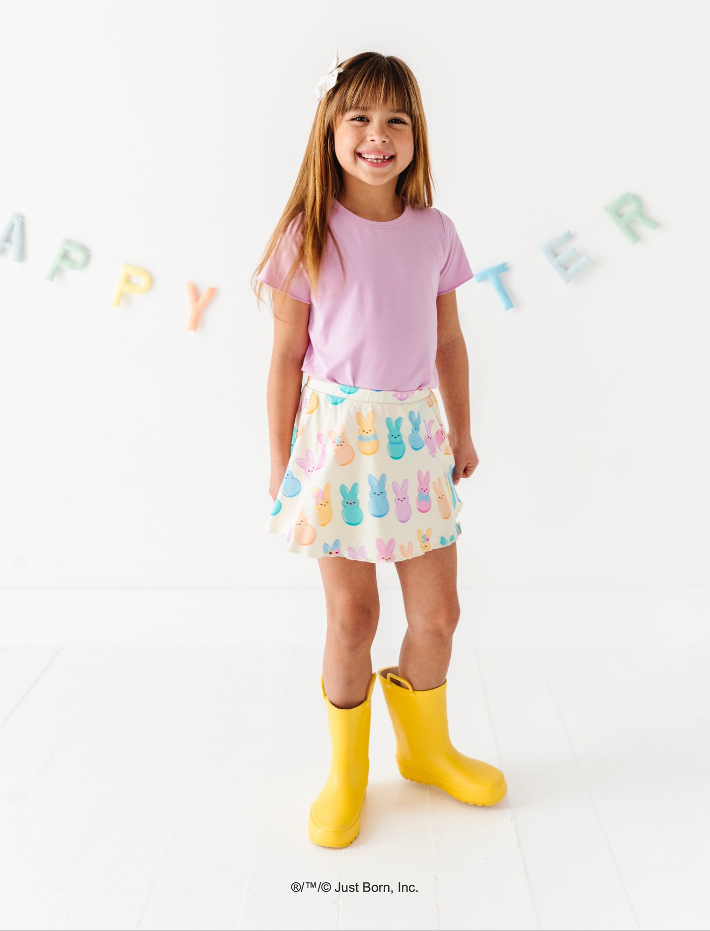 Young girl wearing a pink shirt, patterned PEEPS® bamboo skirt, and yellow rain boots on a white background with 'Happy Easter' text.