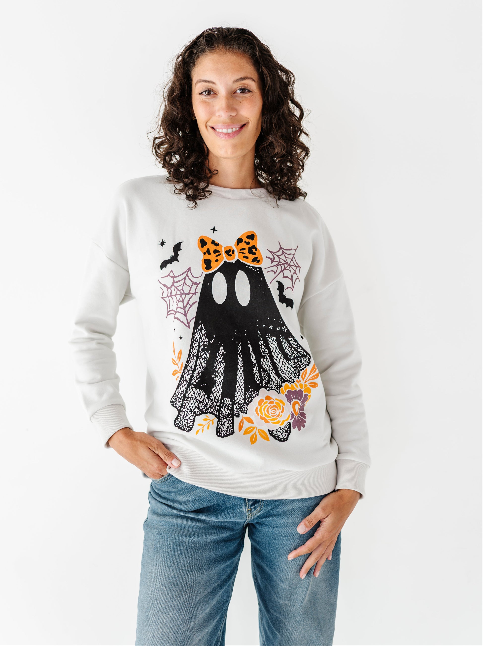 A woman wearing a beige crewneck sweatshirt with a large black ghost graphic decorated with an orange leopard-print bow, black lace, orange roses, and bats, paired with blue jeans against a plain white background.