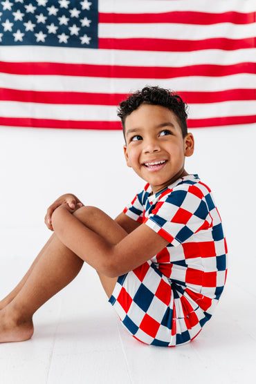 Child wearing a red, white, and blue checkered bamboo pajama outfit sitting in front of an American flag.