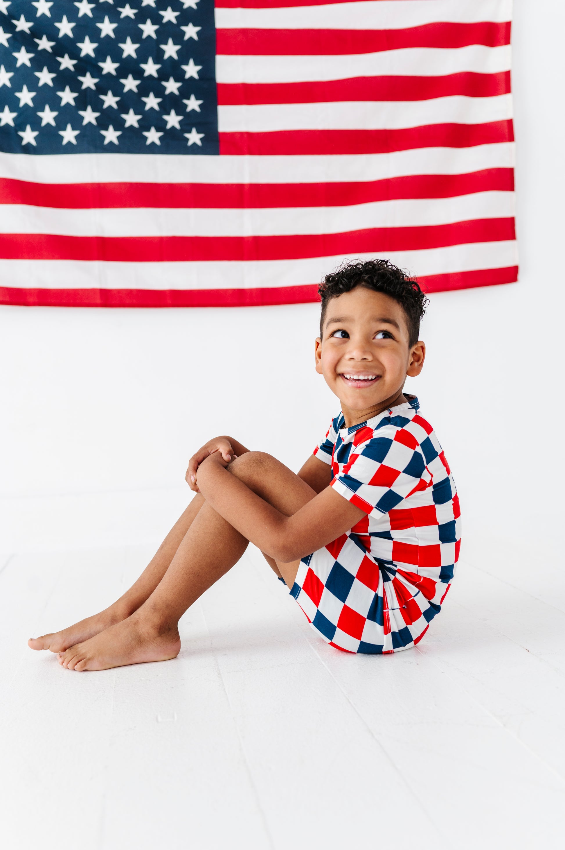 Child wearing a red, white, and blue checkered bamboo short set pajama outfit sitting in front of an American flag.