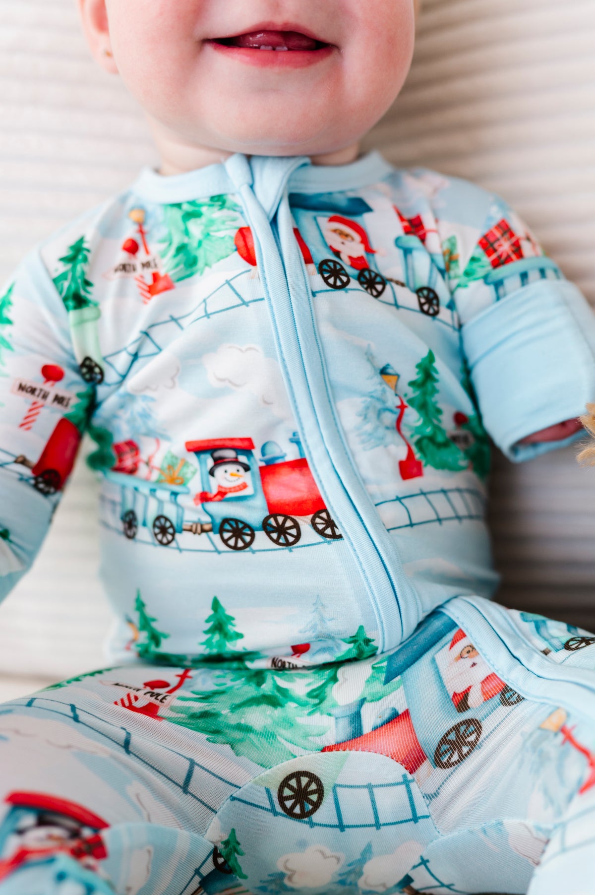 A close-up of a baby wearing a light blue zip-front bamboo sleeper with a festive Christmas train print featuring Santa Claus, snowmen, and pine trees.