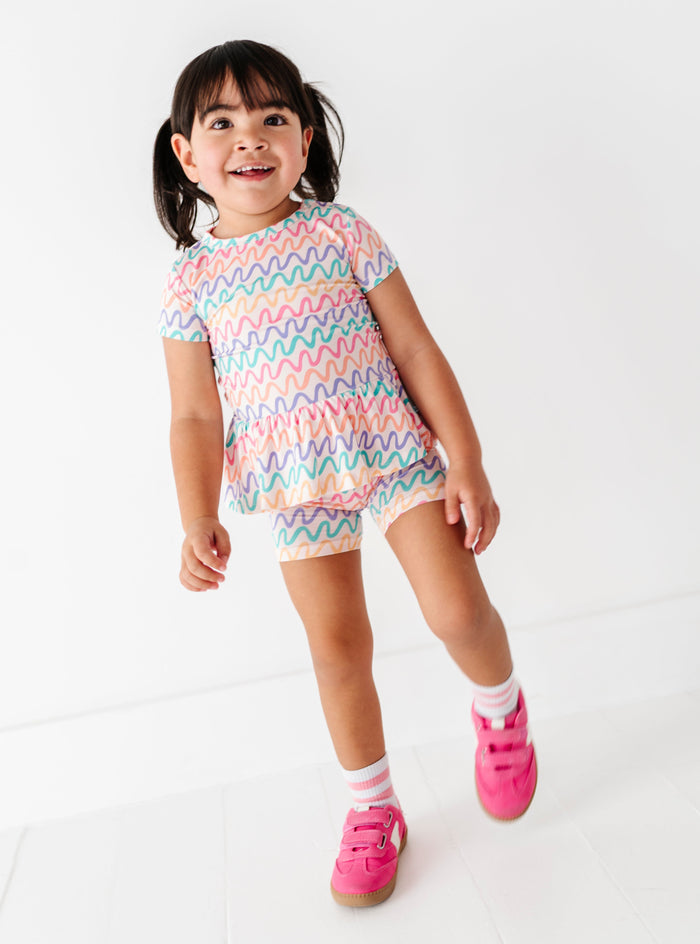 Little girl wearing a colorful squiggly pattern bamboo/cotton peplum top and coordinating biker shorts on a white background