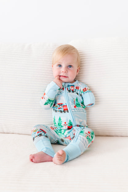 A baby sitting on a cream ribbed cushion wearing a light blue zip-up one-piece bamboo pajama with a Christmas train and tree pattern.