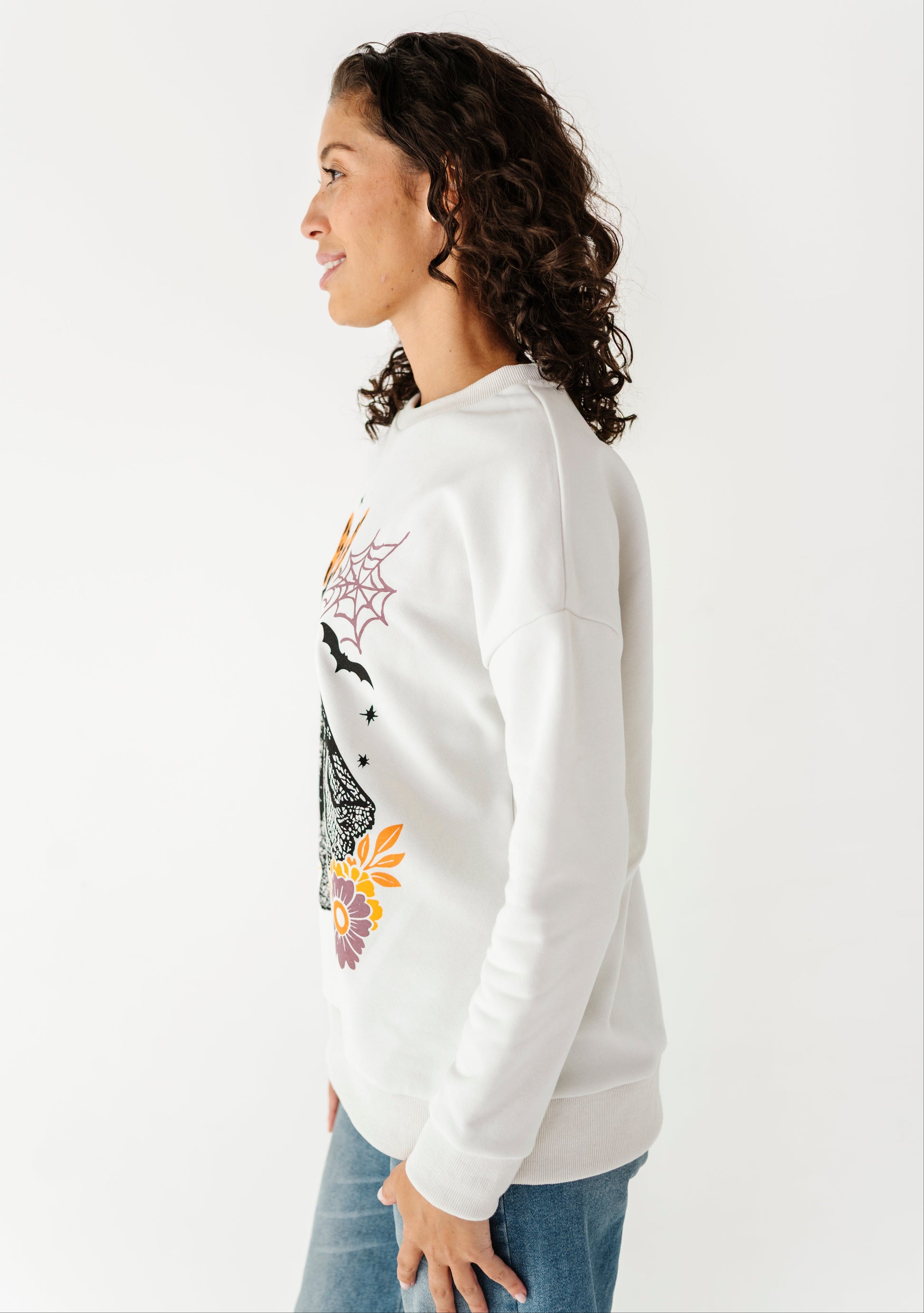A woman in profile wearing a white crewneck sweatshirt with a colorful spiderweb and spider graphic on the front, paired with blue jeans, against a plain white background.