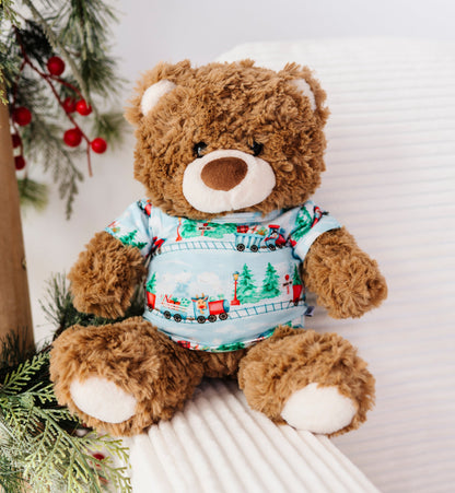 A brown plush teddy bear wearing a light blue Christmas-themed bamboo t-shirt with a train pattern, sitting on a white ribbed chair next to a decorated Christmas tree branch with red berries.