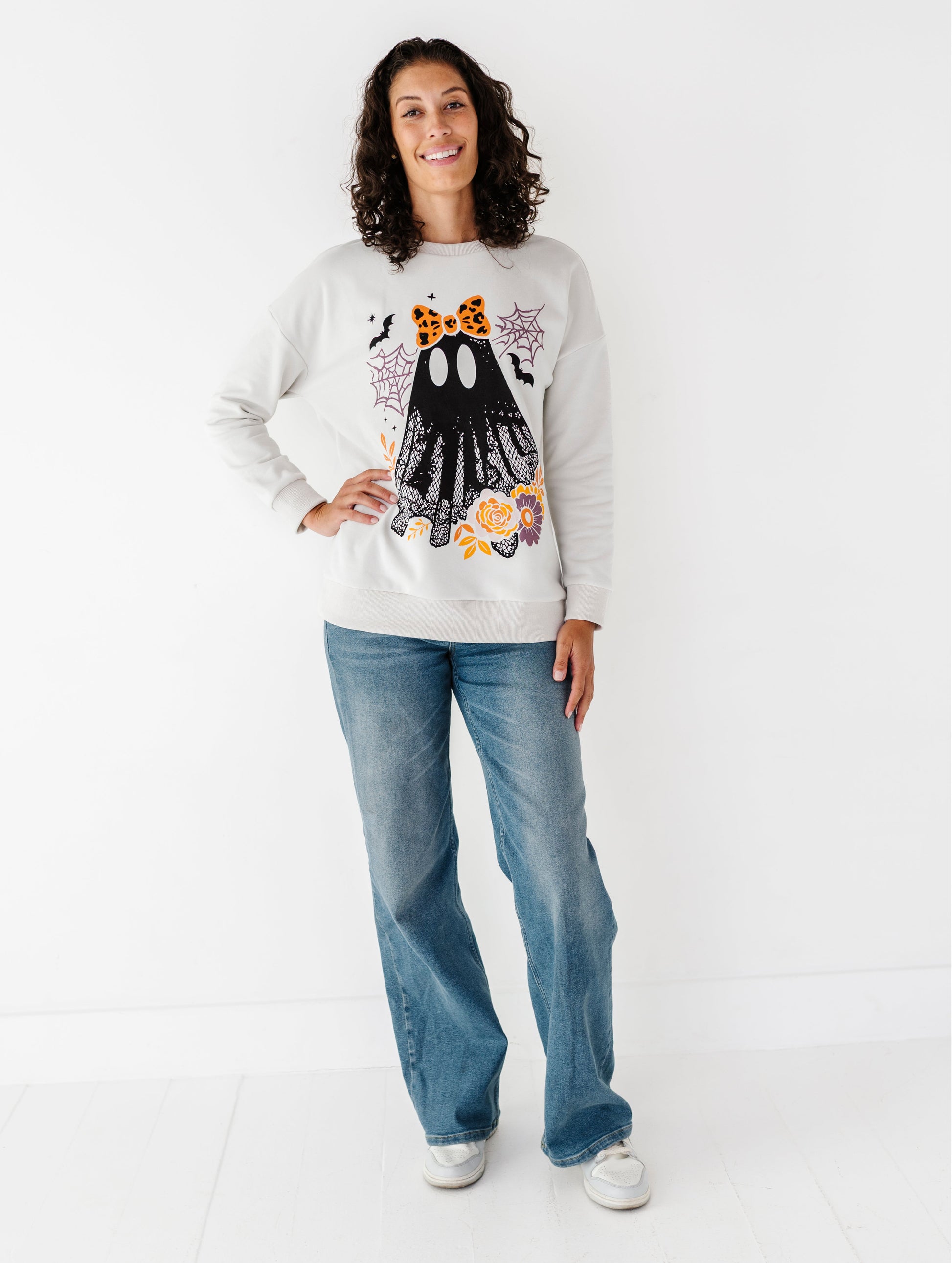 A woman modeling a light gray crewneck sweatshirt with a Halloween-themed ghost graphic, paired with blue jeans and white sneakers, standing against a plain white studio background.