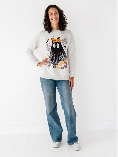 A woman modeling a light gray crewneck sweatshirt with a Halloween-themed ghost graphic, paired with blue jeans and white sneakers, standing against a plain white studio background.