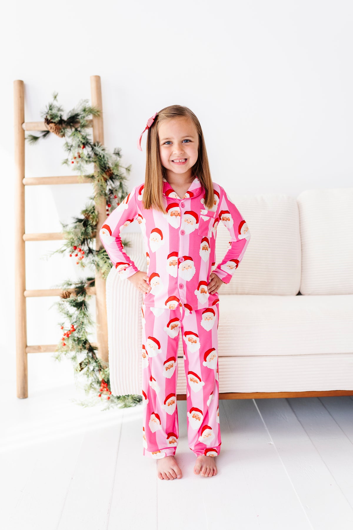 A young girl stands barefoot in a bright, white room wearing a pink Santa Claus patterned bamboo pajama set. A wooden ladder with holiday greenery is on the left, and a white sofa is on the right.