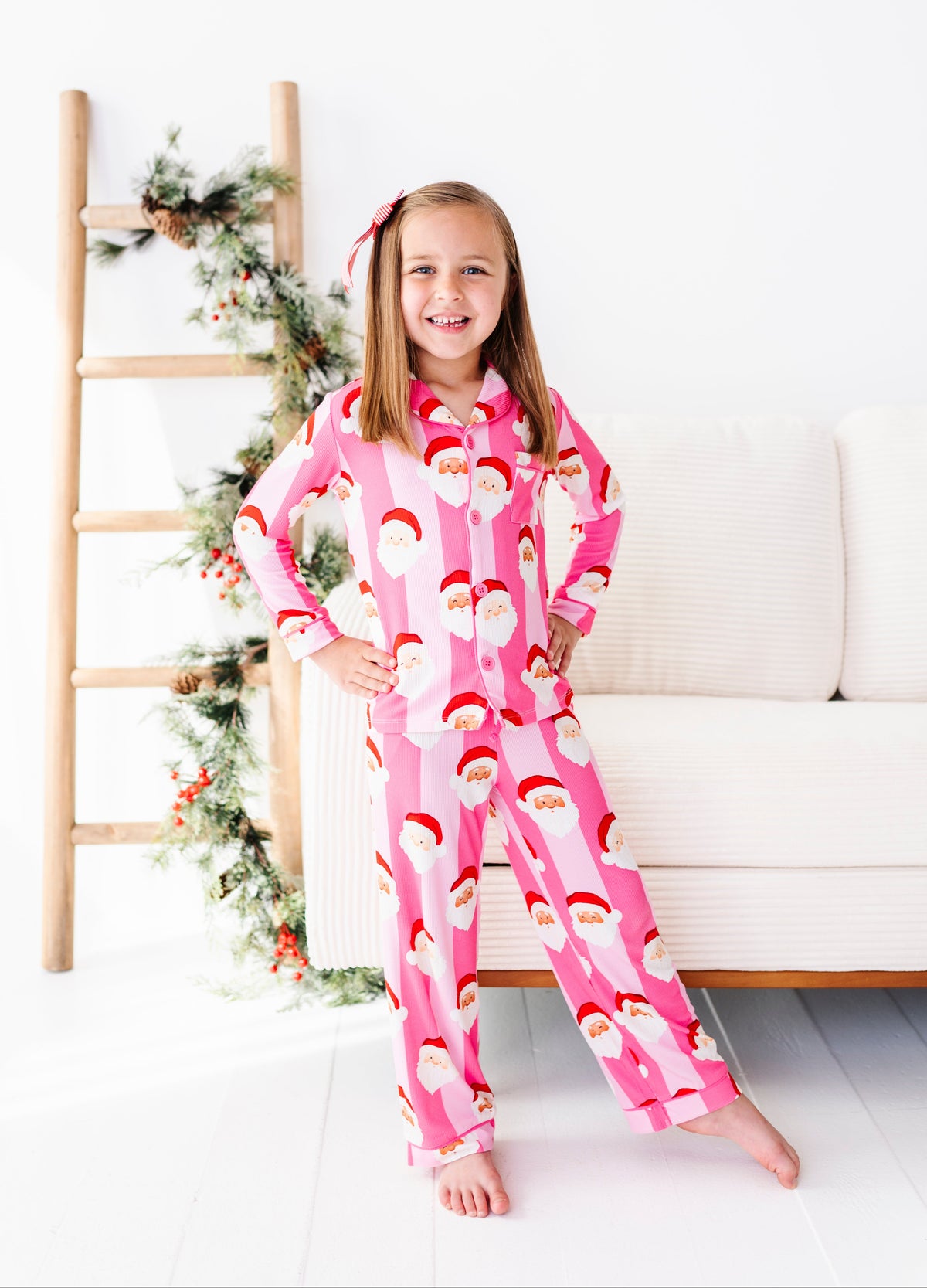 A young girl smiling while wearing a pink Santa Claus patterned bamboo pajama set, standing in a bright room next to a white sofa and a wooden ladder decorated with holiday greenery.