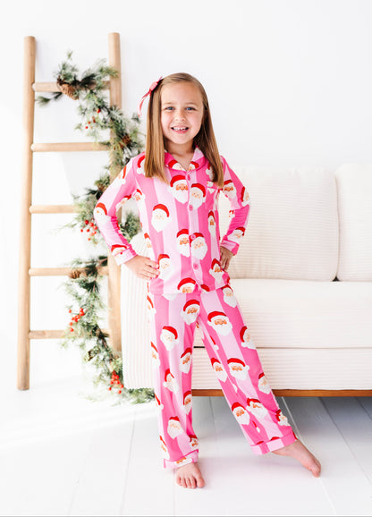 A young girl smiling while wearing a pink Santa Claus patterned bamboo pajama set, standing in a bright room next to a white sofa and a wooden ladder decorated with holiday greenery.