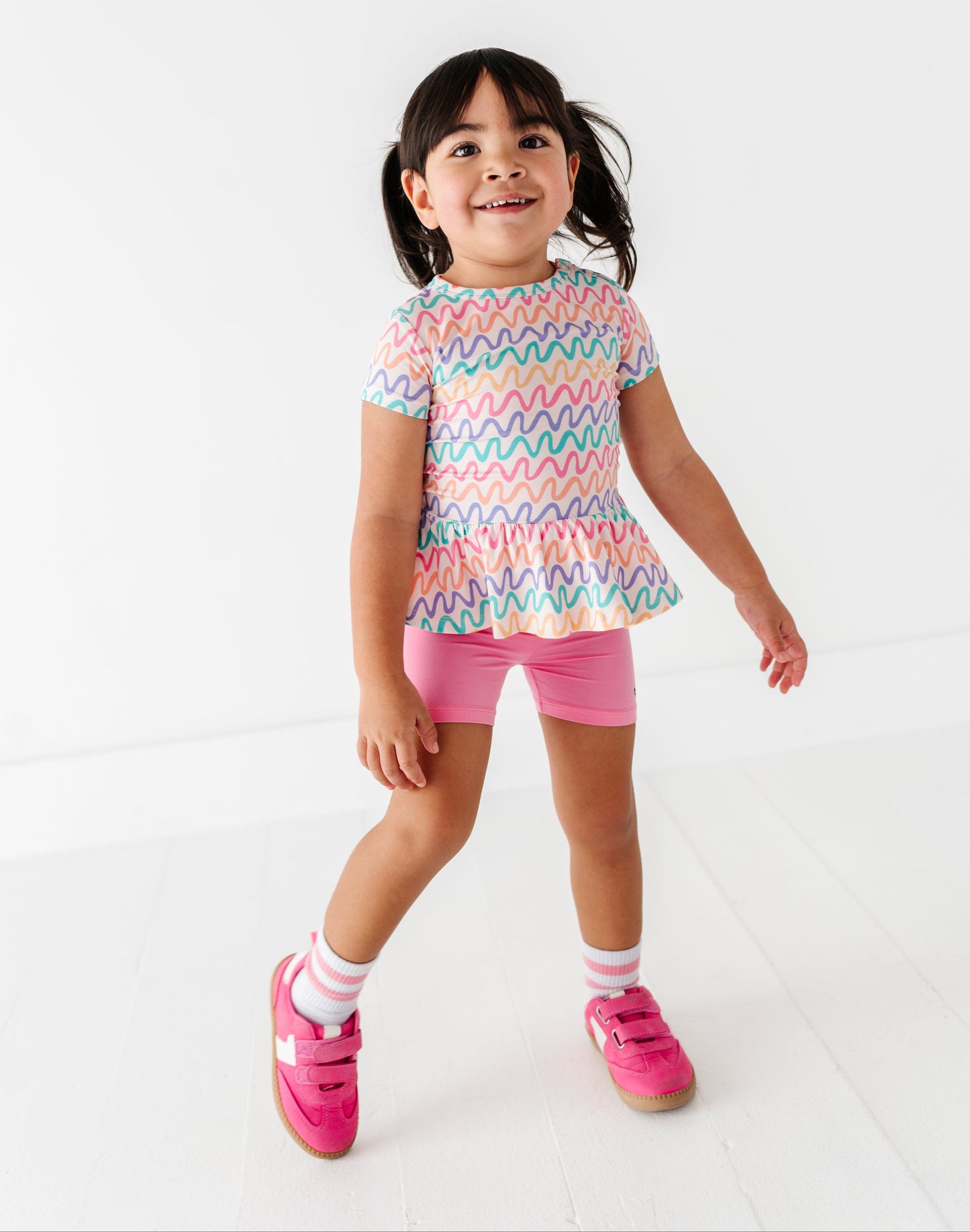 Young girl wearing a colorful wavy patterned bamboo/cotton top and pink bamboo biker shorts on a white background