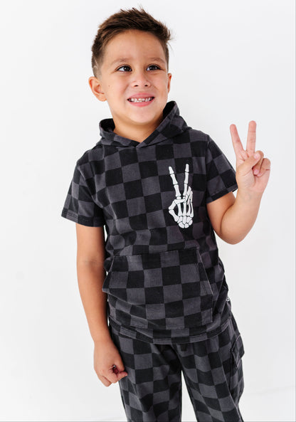 A young boy smiling and posing with a peace sign, wearing a matching black and gray checkered hoodie and sweatpants set with a white skeleton hand graphic on the chest.