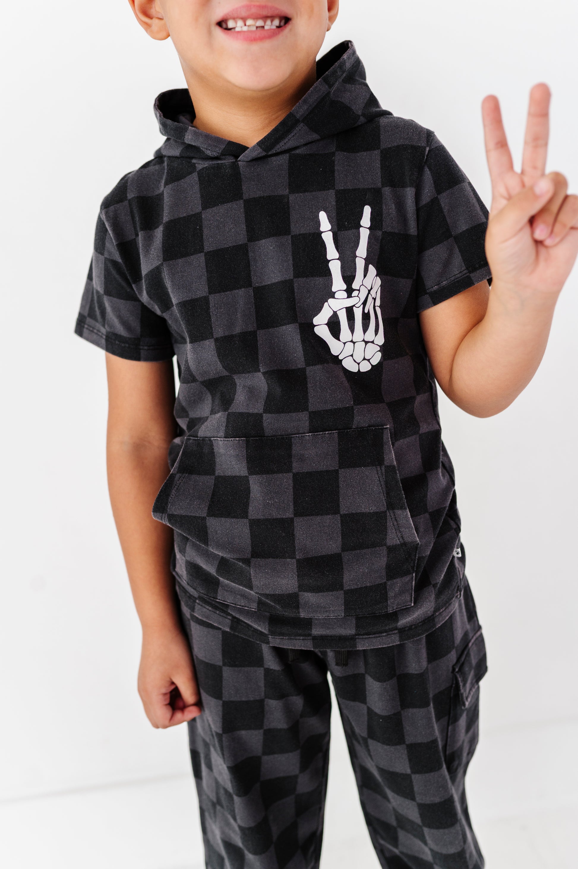 A smiling child wearing a black and dark gray checkered hoodie and matching pants, with a white skeleton hand graphic on the chest, posing against a white background.