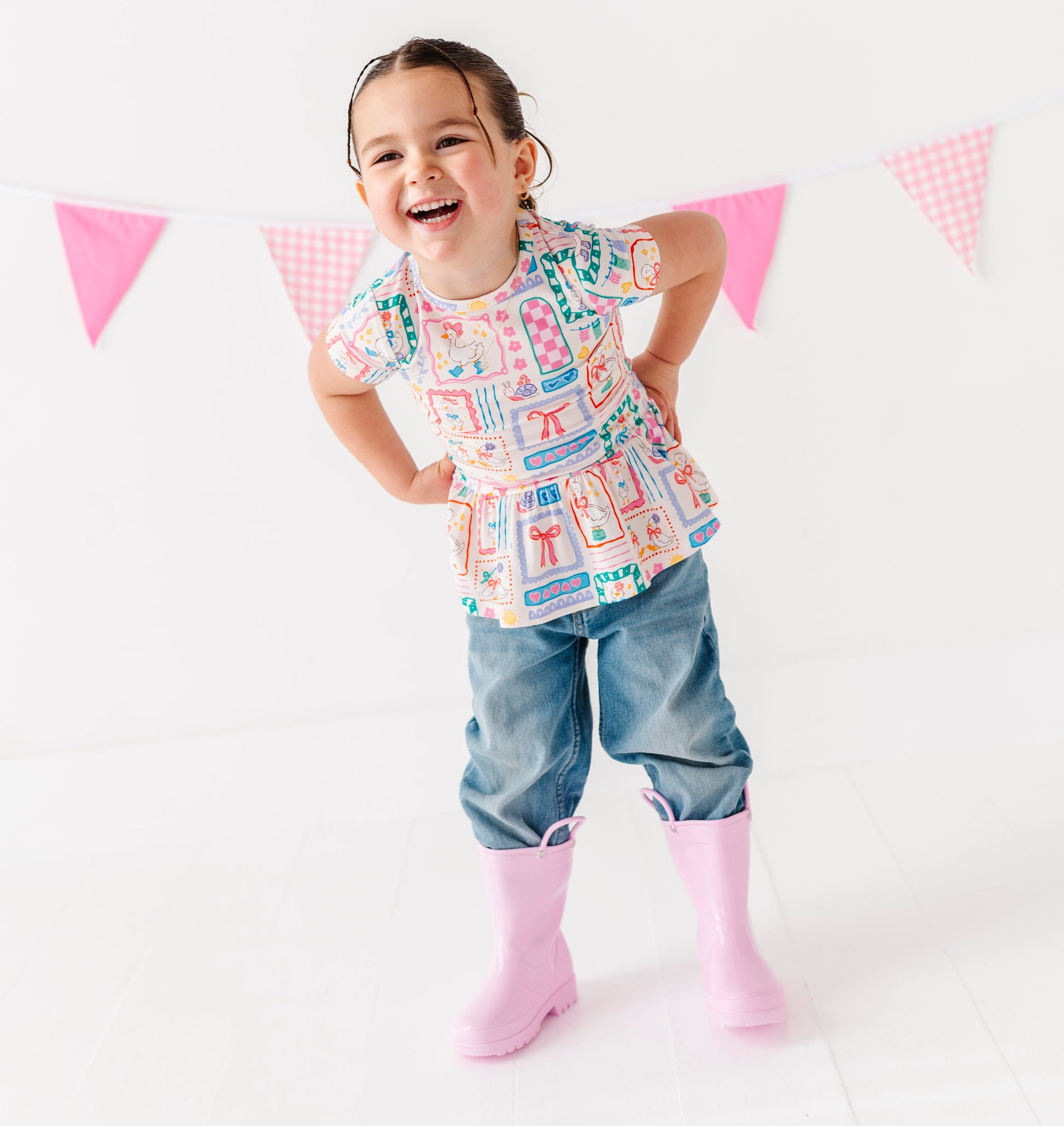 Child wearing a colorful Gilly Geese peplum shirt and denim pants standing in front of pink and white flags on a white background