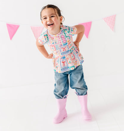 Child wearing a colorful Gilly Geese peplum shirt and denim pants standing in front of pink and white flags on a white background