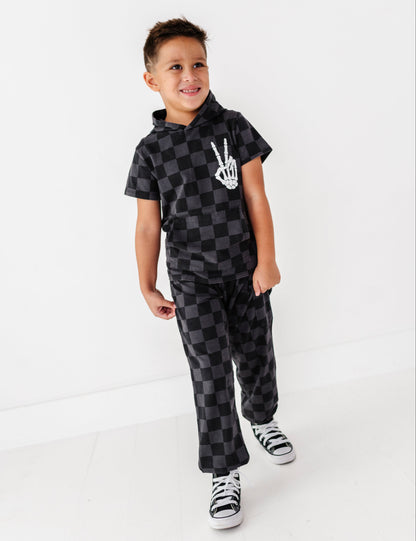 A young boy modeling a black and gray checkered hooded sweatshirt and matching sweatpants with a white skeleton hand graphic on the chest, paired with black and white sneakers, set against a plain white studio background.