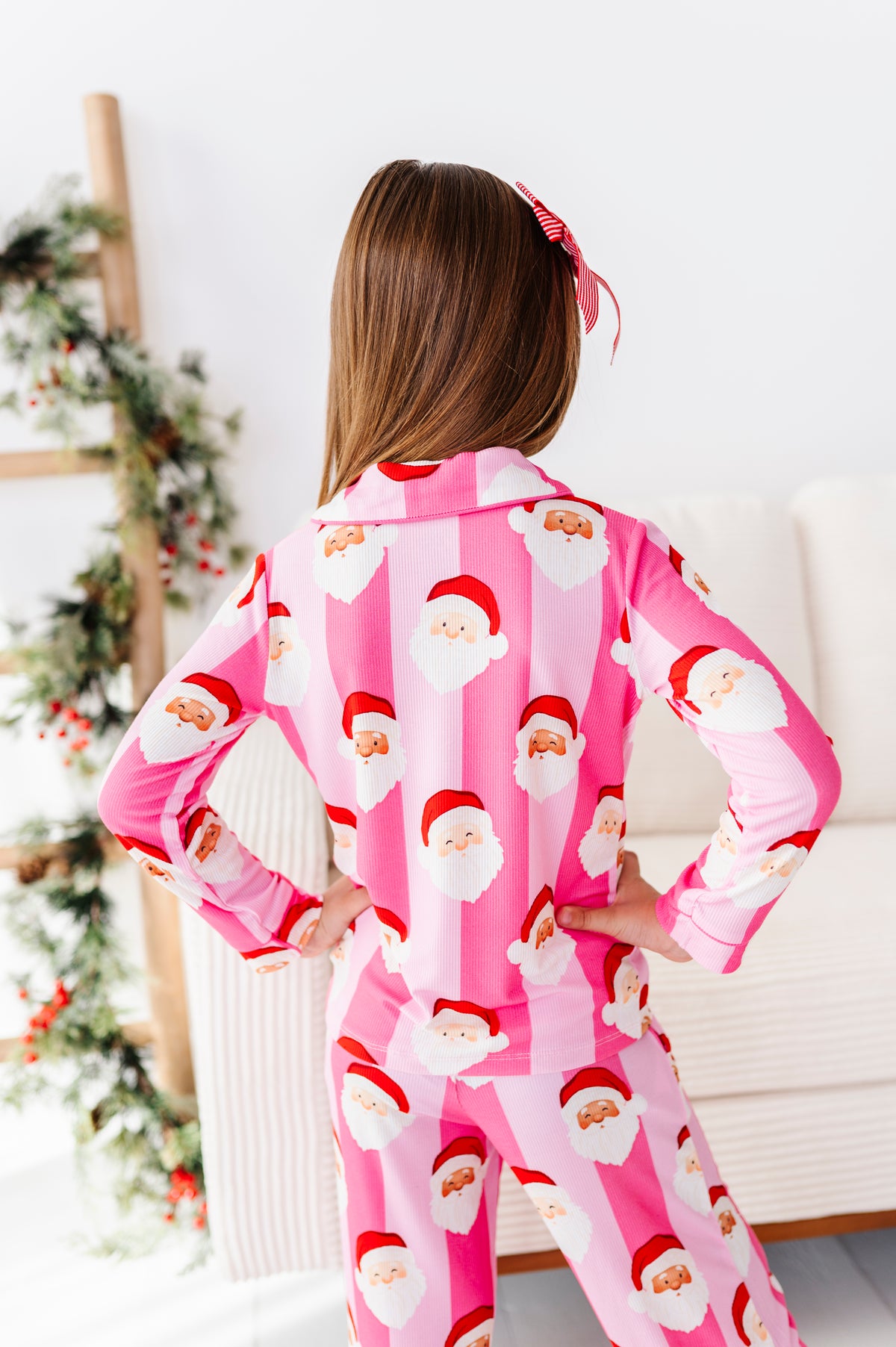 A child wearing a pink Santa Claus bamboo pajama set, shown from the back in a brightly lit room with a holiday wreath and sofa in the background.